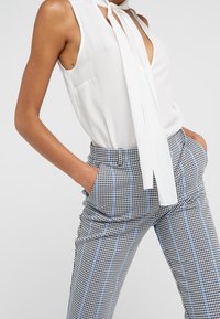 Blusa bianca senza maniche con cravatta, abbinata a pantaloni a quadri aderenti in bianco e nero con accenti blu. Dettaglio distintivo della tasca.