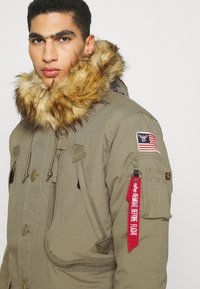 Parka verde militare con colletto in pelliccia sintetica rimovibile, doppie tasche frontali, tasca con zip sulla manica e un'etichetta rossa "Rimuovere prima del volo".