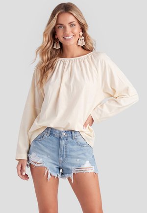 Lächelnde Frau mit langen, welligen blonden Haaren, die eine beige Bluse mit langen Ärmeln, abgetragene blaue Jeansshorts und Quastenohrringe trägt und mit der Hand in der Hüfte steht.