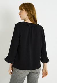 Top negro con escote redondo y cuello con volantes. Presenta mangas largas con puños con volantes. Tela suave y ligera, diseño simple.