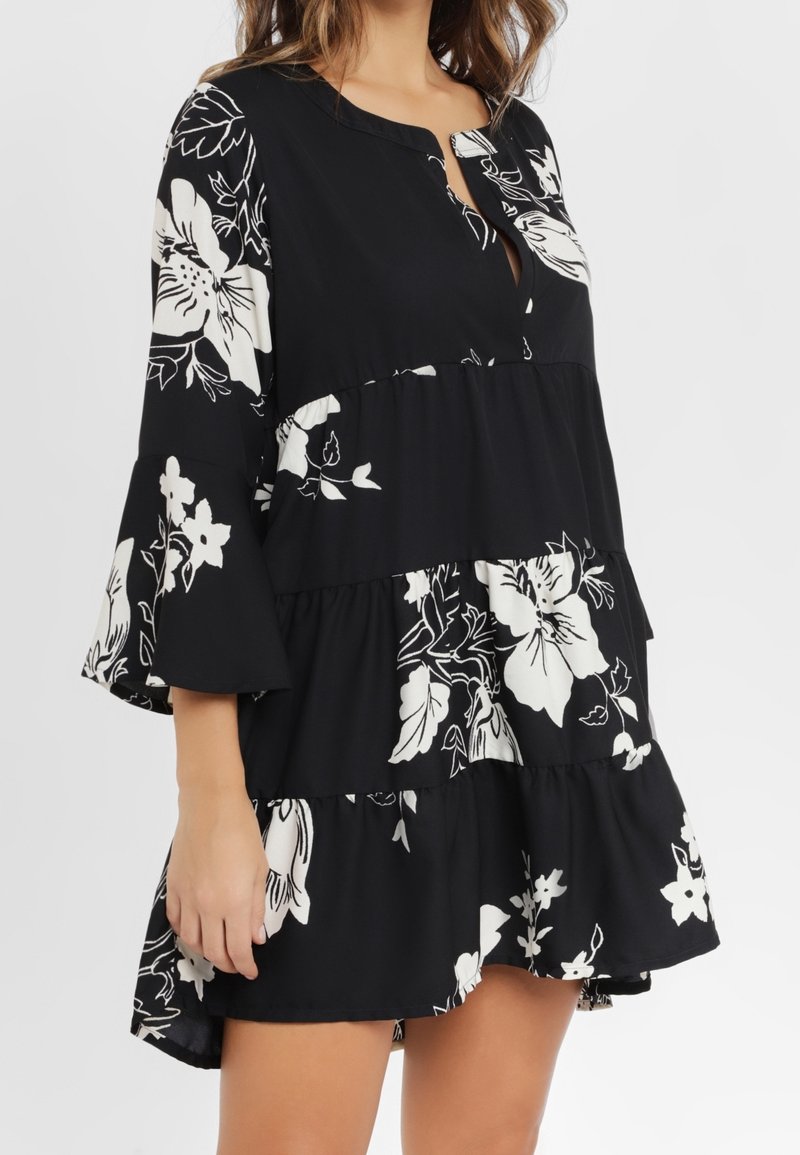 Robe noire à étages avec imprimé floral blanc, manches trois-quarts évasées et encolure ronde avec une petite fente à l’avant.
