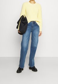 Stickad gul tröja med lätt textur, kombinerad med blå denimjeans, rak passform, bärs med svarta kraftiga stövlar och en svart vadderad väska.