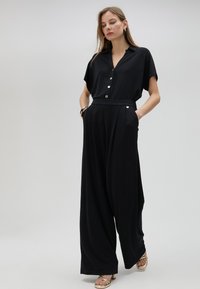 Mono negro de tejido suave, con un frente abotonado, mangas cortas, diseño de pierna ancha y bolsillos laterales. Acentos dorados en la cinturilla.