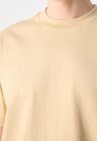 T-shirt beige à col rond avec manches courtes et logo "HUGO" embossé sur la poitrine, porté par une personne visible du cou jusqu'au milieu du torse.