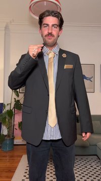 Casaco de fato preto sobre camisa às riscas, gravata dourada e lenço de bolso. A pessoa segura um microfone. Ambiente interior com plantas.