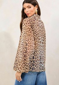 Blouse en dentelle léopard beige et marron, dotée de manches longues et d'un col montant. Portée par-dessus un jean en denim bleu.