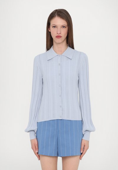 Lichtblauwe gebreide blouse met een puntige kraag, knoopsluiting en geribbelde structuur. Lange mouwen met aansluitende manchetten. Gecombineerd met blauw gestreepte shorts.