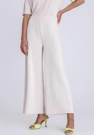 Vrouw die crèmekleurige wijde broek en lichtgroene open-toe sandalen met hak draagt, met één hand op de heup.