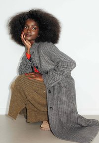 Manteau en pied-de-poule gris et noir avec de grands boutons, superposé à une jupe côtelée marron. Un top rouge et des bottes beiges complètent la tenue.