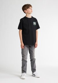 Zwarte katoenen t-shirt met een vierkant logo op de linkerborst, gecombineerd met grijze spijkerbroek en witte sneakers. Eenvoudig en casual ontwerp.