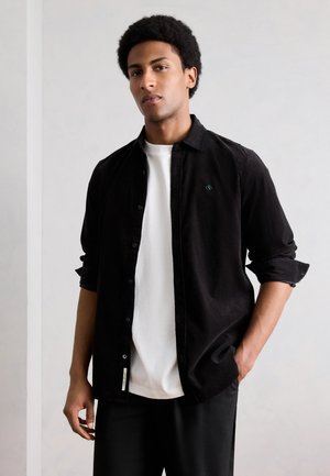 Junger Mann mit Afro, der ein schwarzes Hemd über einem weißen T-Shirt trägt, steht mit einer Hand in der Tasche vor einem schlichten Hintergrund.