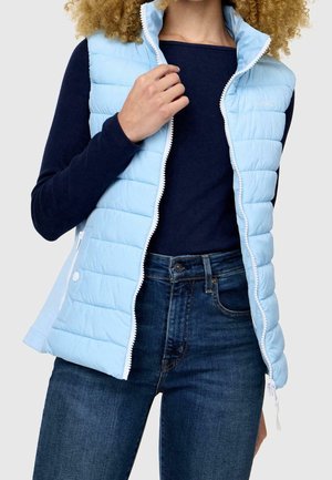 Femme portant un gilet matelassé bleu clair par-dessus un t-shirt manches longues bleu marine et un jean bleu, tenant le col du gilet près de la fermeture éclair sur un fond gris.