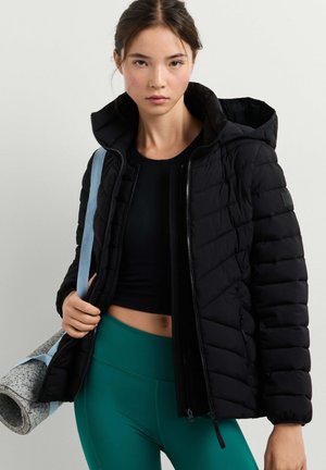 Junge Frau trägt eine schwarze gesteppte Jacke mit Kapuze, ein dunkles Crop-Top und grüne Leggings, hält eine zusammengerollte Yogamatte mit hellblauem Riemen.
