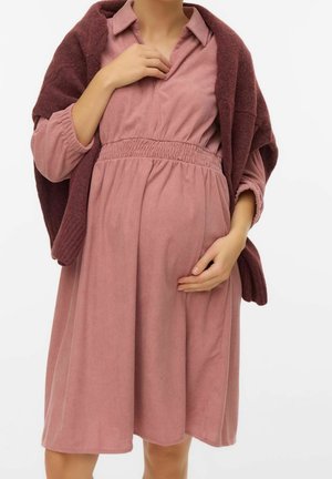 Zwangere persoon draagt een mauve ribfluwelen jurk met een geplooide taille, een bordeauxrode trui over de schouders gedrapeerd, hand rustend op de buik.