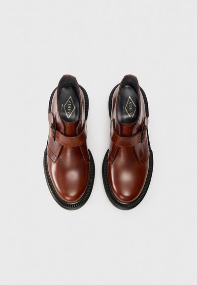Scarpe in pelle marrone con punta arrotondata, dotate di una cinghia con fibbia e una suola in gomma nera testurizzata per aderenza e durata.