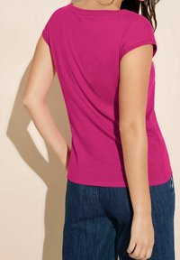 Pěkně padnoucí tričko s krátkým rukávem v jasně růžové barvě z bavlny. Obsahuje klasický kulatý výstřih a jemné detaily šití. V kombinaci s tmavě modrými džíny.