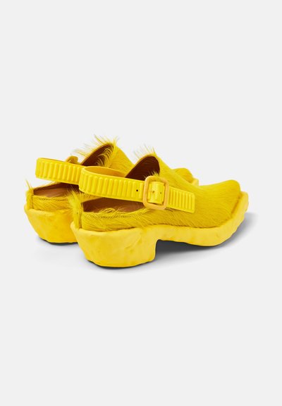 Gelbe, mit Fleece überzogene Clogs mit einer strukturierten Oberfläche, die eine dicke Sohle und einen verstellbaren Plastikverschlussriemen an der Rückseite haben.