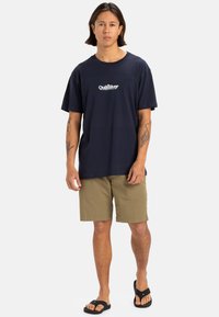Camiseta de algodón azul marino con el logo de Quiksilver, combinada con pantalones cortos color caqui y chanclas negras. Diseño simple, corte relajado, mangas cortas.