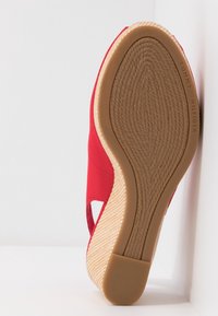 Chaussure compensée en tissu rouge avec une semelle en caoutchouc texturé couleur tan. Présente un design avec une découpe sur le côté et un accent tissé naturel le long de la semelle.