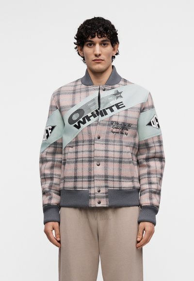 Jeune homme aux cheveux bouclés portant une veste à carreaux rose et grise avec des rayures diagonales bleues et le texte « OFF WHITE », debout devant un fond blanc uni.