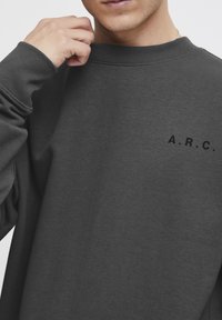 Dark grey sweatshirt with ribbed cuffs and neckline. Features "A.R.C." in black lettering on the front. Fabric appears soft and textured.