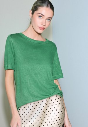 Woman with light skin and blue eyes wearing a green short-sleeve shirt and a beige skirt with black polka dots, standing against a plain background.