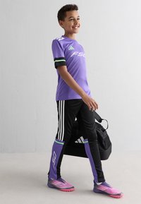 Μακριά μπλούζα αθλητισμού σε μωβ χρώμα με πράσινες και μαύρες λεπτομέρειες, μαύρο παντελόνι με λευκές ρίγες και μωβ παπούτσια με ροζ λεπτομέρειες, μαζί με μια μαύρη σάκα γυμναστικής.