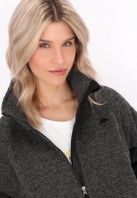 Sivi fleece jakna na zatvarač s visokim ovratnikom, s blagom teksturom i malim logom s lijeve strane. Ispod nje je vidljiva bijela košulja.