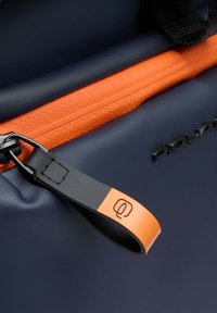 Détail d'un sac imperméable marine avec une fermeture éclair texturée orange et une languette noire avec un petit accent orange sur son bord.