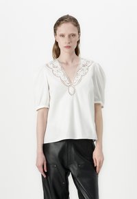 Blouse en coton blanche avec des manches bouffantes et un décolleté en V profond, ornée de détails en dentelle délicate autour du col. Portée avec un pantalon en cuir noir.