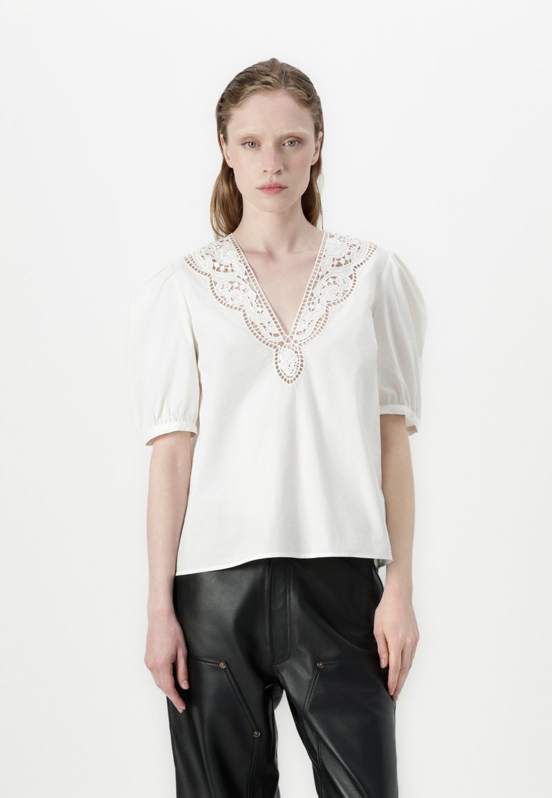 Blouse en coton blanche avec des manches bouffantes et un décolleté en V profond, ornée de détails en dentelle délicate autour du col. Portée avec un pantalon en cuir noir.