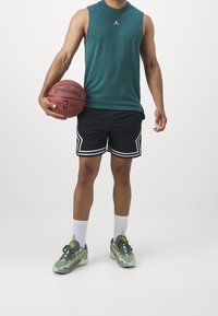 Camiseta verde sin mangas, pantalones cortos deportivos negros con ribete blanco, calcetines blancos y zapatillas verdes sosteniendo un balón de baloncesto Spalding.