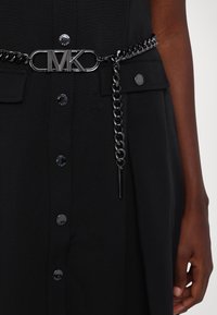 Ceinture en chaîne noire avec une boucle en métal rectangulaire portant le logo "MK", attachée à un tissu texturé avec des accents de boutons métalliques.