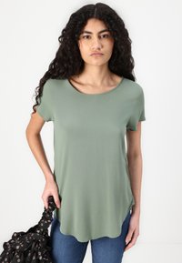 Olive green short-sleeve top with a round neckline and a slightly curved hem. Model holds a black patterned bag in her right hand.