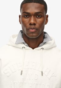 Hvid hoodie med "SUPERDRY N.Y.C." præget på forsiden. Har en kontrasterende stribet krave og snorede detaljer. Glat tekstur.