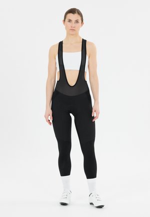 Athletische vrouw die een witte sportbeha, zwarte fietsbroek met bretels, witte sokken en witte fietsschoenen draagt, staand tegen een effen witte achtergrond.