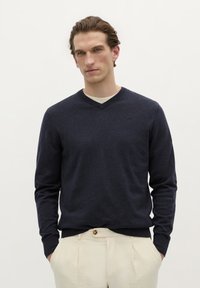 Marineblauer V-Ausschnitt-Pullover aus weichem Stoff, mit langen Ärmeln und taillierter Passform. Über einem hellbeigen Hemd getragen.