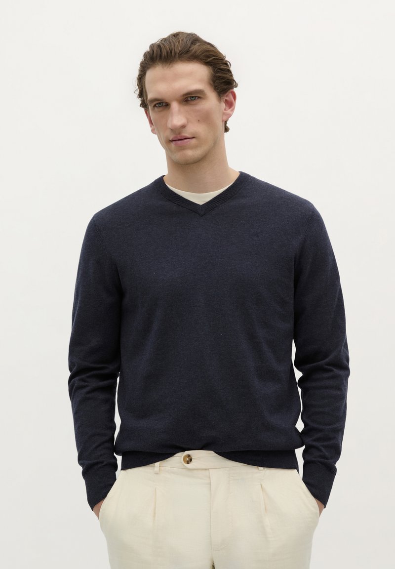 Marineblauer V-Ausschnitt-Pullover aus weichem Stoff, mit langen Ärmeln und taillierter Passform. Über einem hellbeigen Hemd getragen.