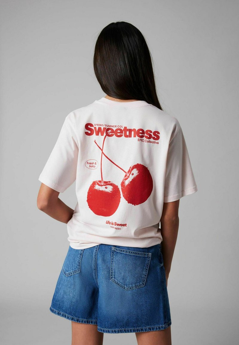 Witte t-shirt met korte mouwen met een rode kersafbeelding op de achterkant, met de tekst "Zoetheid" en "Het leven is zoet" eronder gedrukt.