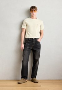 Világos bézs színű póló kis grafikai mintával, sötét farmernadrággal és barna slip-on cipővel. A modellen túlméretezett narancsszínű napszemüveg van.