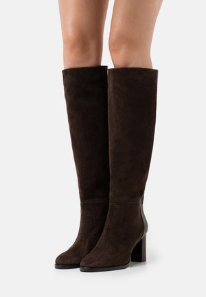 Stivali scamosciati marrone scuro al ginocchio con tacchi a blocco, indossati su gambe nude contro uno sfondo chiaro uniforme.