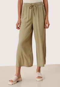 Jasnozielone spodnie o szerokich nogawkach wykonane z gładkiej tkaniny, z elastycznym pasem i sznurkiem do regulacji. Do tego beżowe, masywne klapki.