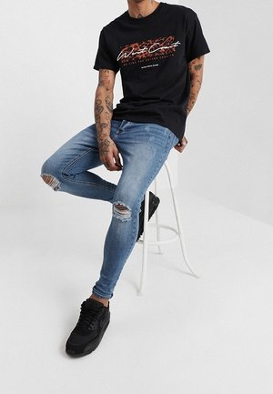 Mann trägt schwarzes T-Shirt mit Grafik, zerrissene enge blaue Jeans und schwarze Turnschuhe, sitzt auf einem weißen Hocker vor einfarbigem Hintergrund.