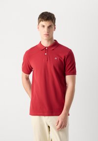 Rotes Polo-Shirt mit klassischem Kragen und kurzen Ärmeln, versehen mit einem kleinen Logo auf der linken Brust. Weiches Baumwollmaterial mit glatter Textur.