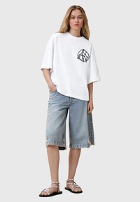 T-shirt bianca oversize con grafica nera sul petto, abbinata a pantaloncini di denim chiaro strappati e sandali argentati con lacci.