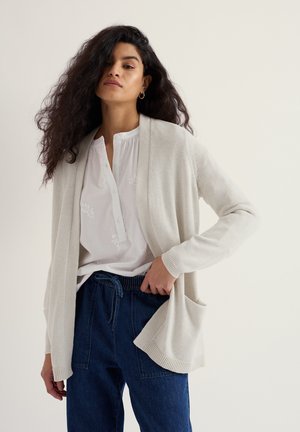 Frau mit langen lockigen Haaren, die eine cremefarbene Strickjacke, eine weiße bestickte Bluse und dunkelblaue Hose mit Kordelzug vor einem einfarbigen Hintergrund trägt.