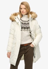 Hvid pufferjakke med en pelsforet hætte, frontlommer og lynlås, båret over en strikket mønstrede sweater og brune bukser.