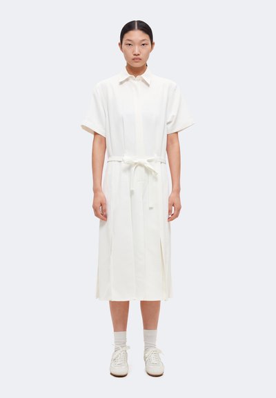 Femme aux cheveux attachés portant une robe midi blanche ceinturée à manches courtes et des baskets blanches, debout devant un fond uni.