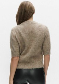 Brun kortärmad tröja gjord av mjukt, luddigt material. Har en rund halsringning och en något kortare fåll. Svarta åtsittande byxor delvis synliga.