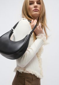 Μαύρη δερμάτινη τσάντα χειρός με καμπυλωτό σχήμα και έναν ιμάντα, με ασημί μεταλλικά στοιχεία, κρατημένη πάνω σε λευκό ριμπ τοπ με δαντελένιο τελείωμα.